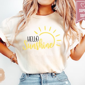 Puede incluir: Camiseta color marfil con las palabras "Hello Sunshine" en negro y amarillo, con un gr&aacute;fico de sol amarillo. La camiseta de manga corta est&aacute; hecha de un material suave. Visible una pancarta rosa con "Super Soft".