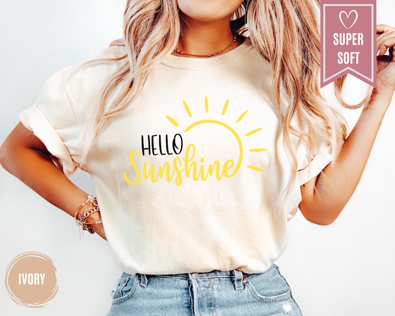 Puede incluir: Camiseta color marfil con las palabras "Hello Sunshine" en negro y amarillo, con un gr&aacute;fico de sol amarillo. La camiseta de manga corta est&aacute; hecha de un material suave. Visible una pancarta rosa con "Super Soft".
