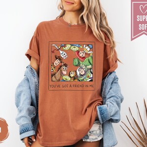 Op de afbeelding: Roestkleurig T-shirt met een ingelijste afbeelding van Toy Story-personages. De afbeelding bevat Woody, Buzz Lightyear en andere personages. De tekst "YOU'VE GOT A FRIEND IN ME" staat eronder. Het shirt heeft opgerolde mouwen.