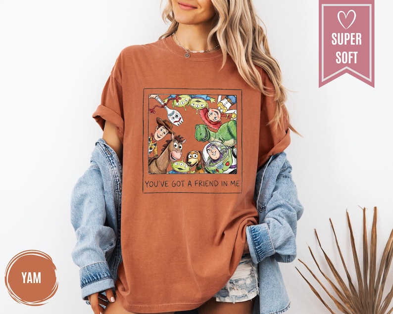 Op de afbeelding: Roestkleurig T-shirt met een ingelijste afbeelding van Toy Story-personages. De afbeelding bevat Woody, Buzz Lightyear en andere personages. De tekst "YOU'VE GOT A FRIEND IN ME" staat eronder. Het shirt heeft opgerolde mouwen.