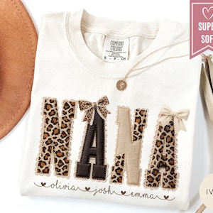 May include: Ivory-colored t-shirt with the word "NANA" in a leopard print design, accented with bows. The names "olivia", "josh", and "emma" are embroidered below. A gold necklace and a hat are visible.