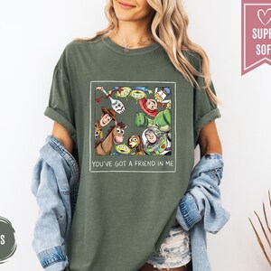 Op de afbeelding: Mosgroen t-shirt met een omlijnde afbeelding van Toy Story-personages. De afbeelding bevat Woody, Buzz Lightyear en de Little Green Men. De tekst "YOU'VE GOT A FRIEND IN ME" staat eronder. Het shirt is gelabeld "SUPER SOFT".