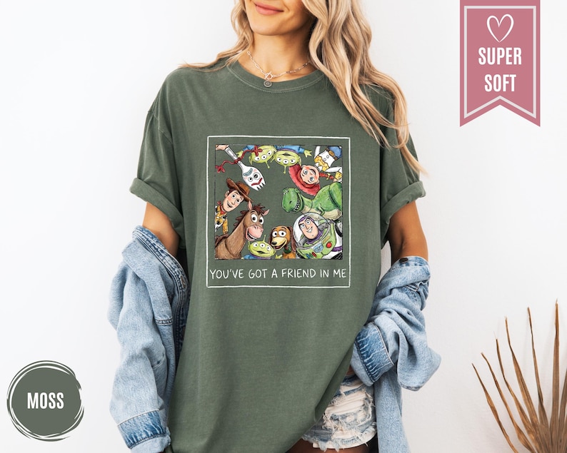 Op de afbeelding: Mosgroen t-shirt met een omlijnde afbeelding van Toy Story-personages. De afbeelding bevat Woody, Buzz Lightyear en de Little Green Men. De tekst "YOU'VE GOT A FRIEND IN ME" staat eronder. Het shirt is gelabeld "SUPER SOFT".