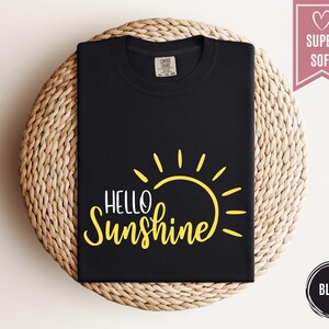 Puede incluir: Camiseta negra con un gr&aacute;fico de sol amarillo y las palabras "Hello Sunshine". La camiseta est&aacute; doblada y se muestra sobre una superficie circular tejida. Una pancarta rosa dice "Super Soft". Un c&iacute;rculo negro dice "Black".