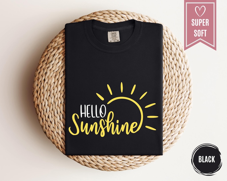 Puede incluir: Camiseta negra con un gr&aacute;fico de sol amarillo y las palabras "Hello Sunshine". La camiseta est&aacute; doblada y se muestra sobre una superficie circular tejida. Una pancarta rosa dice "Super Soft". Un c&iacute;rculo negro dice "Black".