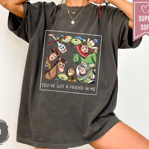 Op de afbeelding: Donkergrijs t-shirt met een kleurrijke afbeelding van personages uit de animatiefilm "Toy Story". De afbeelding is omlijst met de tekst "YOU'VE GOT A FRIEND IN ME" in het wit. Het shirt heeft een relaxte pasvorm en korte mouwen.