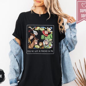 Op de afbeelding: Zwart T-shirt met een kleurrijke afbeelding van personages uit de animatiefilm Toy Story. De afbeelding bevat Woody, Buzz Lightyear en andere personages. De tekst "YOU'VE GOT A FRIEND IN ME" staat onder de afbeelding.