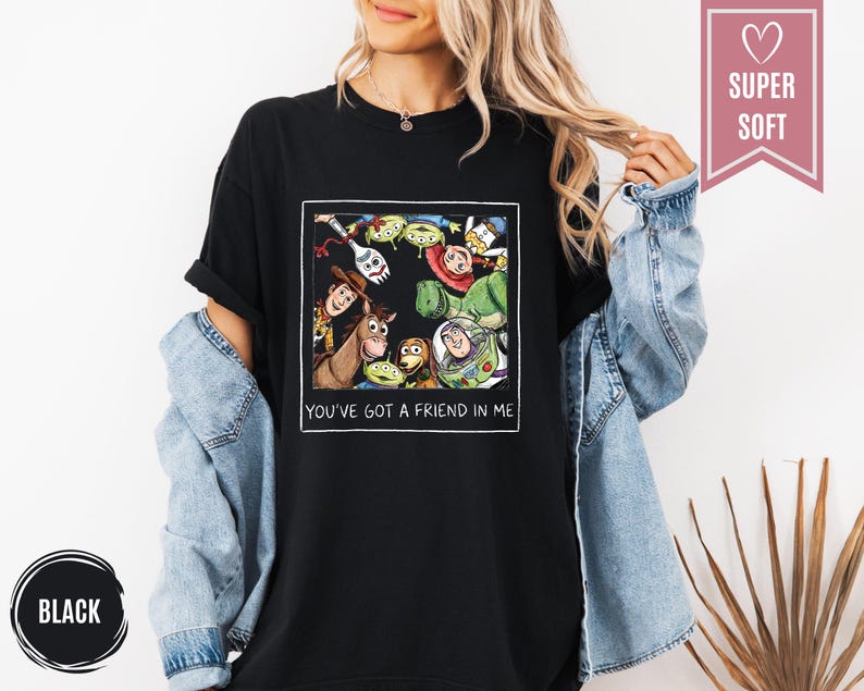 Op de afbeelding: Zwart T-shirt met een kleurrijke afbeelding van personages uit de animatiefilm Toy Story. De afbeelding bevat Woody, Buzz Lightyear en andere personages. De tekst "YOU'VE GOT A FRIEND IN ME" staat onder de afbeelding.