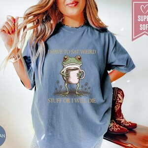 May include: Blue jean colored t-shirt with a graphic of a frog holding a coffee cup. The text on the shirt reads "I HAVE TO SAY WEIRD STUFF OR I WILL DIE". The shirt has a relaxed fit.