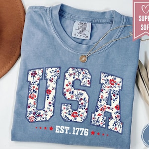 May include: Blue jean colored t-shirt with the word "USA" in a floral pattern. The text "EST. 1776" is below the word "USA". A gold necklace is draped across the shirt. The shirt is labeled "Super Soft".