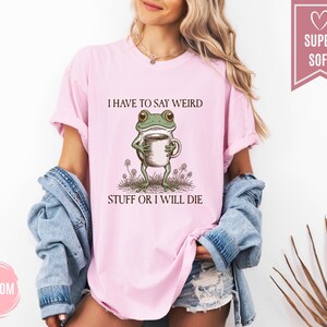 May include: Light pink t-shirt with the text "I HAVE TO SAY WEIRD STUFF OR I WILL DIE" and a cartoon frog holding a coffee cup. The shirt has a relaxed fit and short sleeves. A "SUPER SOFT" tag is visible.