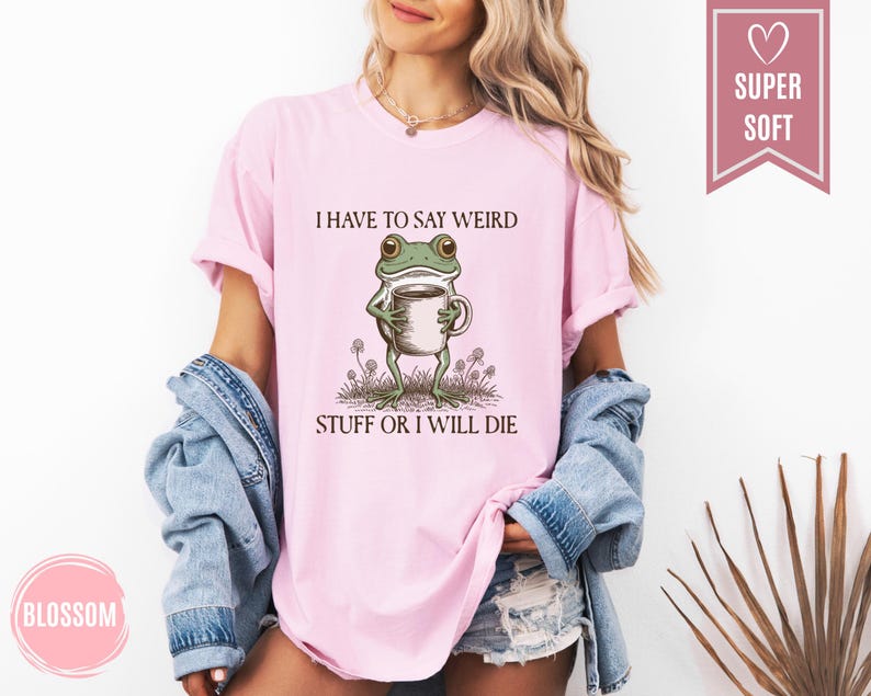 May include: Light pink t-shirt with the text "I HAVE TO SAY WEIRD STUFF OR I WILL DIE" and a cartoon frog holding a coffee cup. The shirt has a relaxed fit and short sleeves. A "SUPER SOFT" tag is visible.