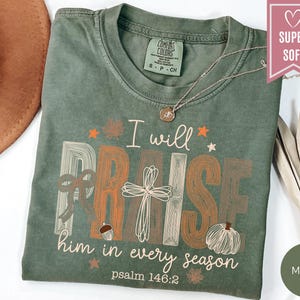 May include: A moss green t-shirt featuring the text I will PRAISE him in every season psalm 146:2. The graphic includes a cross, a bow, an acorn, a pumpkin, and small stars. The shirt has a crew neckline and a Comfort Colors brand label.