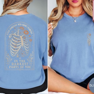 May include: A blue jean colored t-shirt featuring a graphic on the back of a human ribcage surrounded by flowers with the text "FIND SOMEONE WHO GROWS FLOWERS IN THE DARKEST PARTS OF YOU" and a small matching floral graphic on the front chest.