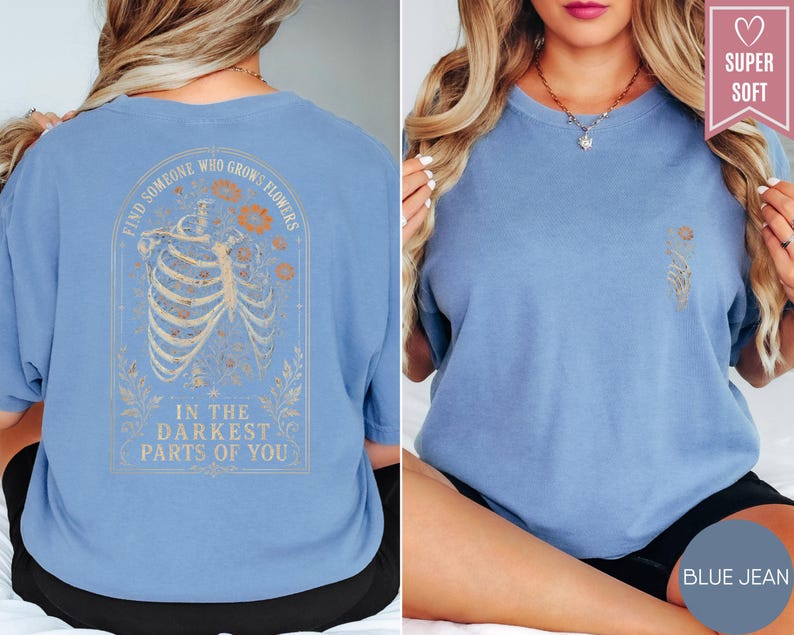 May include: A blue jean colored t-shirt featuring a graphic on the back of a human ribcage surrounded by flowers with the text "FIND SOMEONE WHO GROWS FLOWERS IN THE DARKEST PARTS OF YOU" and a small matching floral graphic on the front chest.