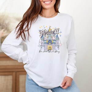 May include: White long-sleeve shirt with a graphic of a castle and the text "Florida!! One Hell Of A Trip".