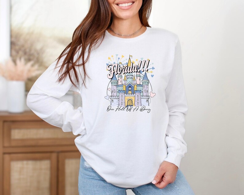 May include: White long-sleeve shirt with a graphic of a castle and the text "Florida!! One Hell Of A Trip".