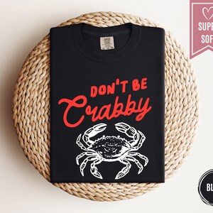May include: Black t-shirt with the text "DON'T BE Crabby" in red script above a white crab illustration. The shirt is folded and displayed on a woven, circular surface. A pink banner with a heart and the words "SUPER SOFT" is on the right.
