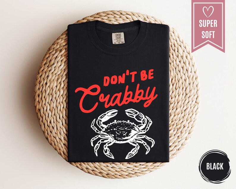 May include: Black t-shirt with the text "DON'T BE Crabby" in red script above a white crab illustration. The shirt is folded and displayed on a woven, circular surface. A pink banner with a heart and the words "SUPER SOFT" is on the right.