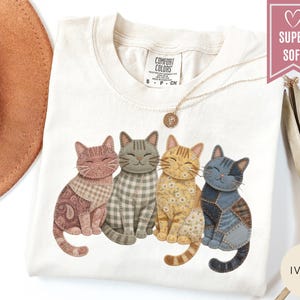 May include: Ivory-colored t-shirt featuring a graphic of four patchwork cats in various colors and patterns. The cats are sitting side-by-side. The shirt has a crew neck and short sleeves. A gold necklace with a pendant is draped on the shirt.