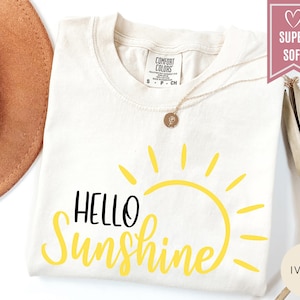 Puede incluir: Camiseta de color marfil con las palabras "HELLO Sunshine" en negro y amarillo. El dise&ntilde;o presenta un gr&aacute;fico de sol amarillo. Un collar de oro con un colgante est&aacute; drapeado sobre la camiseta. La camiseta est&aacute; etiquetada como "SUPER SOFT".