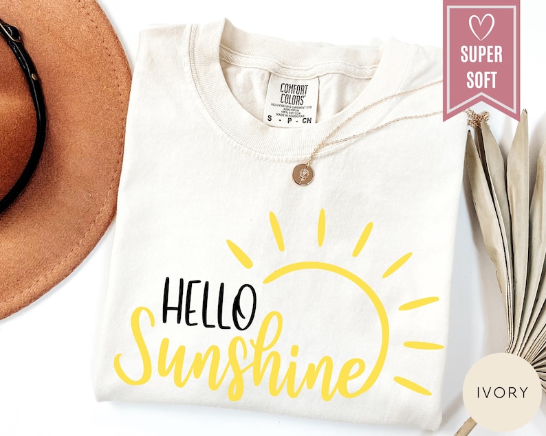 Puede incluir: Camiseta de color marfil con las palabras "HELLO Sunshine" en negro y amarillo. El dise&ntilde;o presenta un gr&aacute;fico de sol amarillo. Un collar de oro con un colgante est&aacute; drapeado sobre la camiseta. La camiseta est&aacute; etiquetada como "SUPER SOFT".