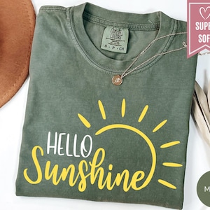 Puede incluir: Una camiseta verde musgo con las palabras "HELLO Sunshine" en escritura amarilla y un gr&aacute;fico de sol amarillo. La camiseta tiene cuello redondo y mangas cortas. Un collar dorado con un colgante est&aacute; en la camiseta.