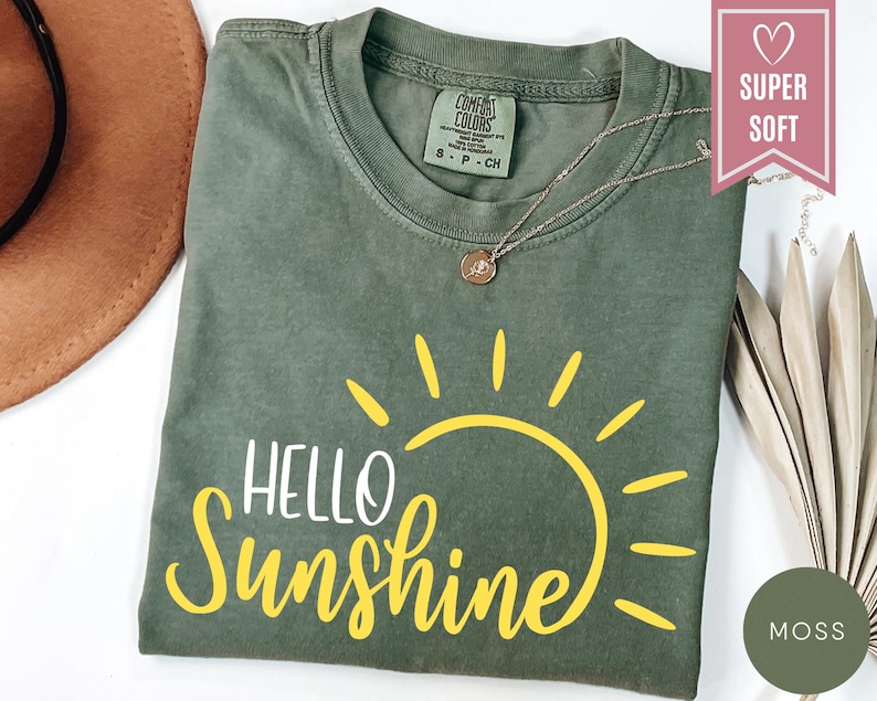 Puede incluir: Una camiseta verde musgo con las palabras "HELLO Sunshine" en escritura amarilla y un gr&aacute;fico de sol amarillo. La camiseta tiene cuello redondo y mangas cortas. Un collar dorado con un colgante est&aacute; en la camiseta.