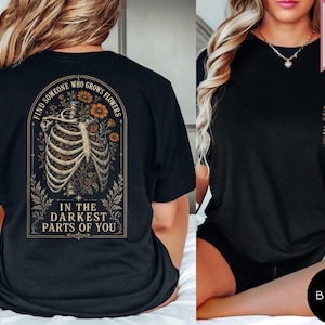 May include: A black t-shirt featuring a graphic on the back of a human ribcage intertwined with orange flowers, framed by the text "FIND SOMEONE WHO GROWS FLOWERS IN THE DARKEST PARTS OF YOU." The front chest area displays a small skeletal hand holding flowers.