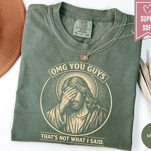 May include: A moss green short-sleeve t-shirt featuring a graphic of a figure with a halo covering their face with one hand. The text reads "OMG YOU GUYS" above the graphic and "THAT'S NOT WHAT I SAID." below it. The shirt has a crew neckline.