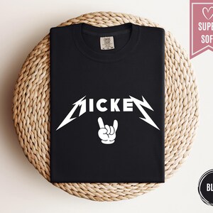 Pu&ograve; includere: T-shirt nera con la scritta "MICKEY" in bianco, con un carattere stilizzato, e un gesto della mano bianco. La maglietta &egrave; piegata su una superficie circolare intrecciata. Uno striscione rosa con un cuore e le parole "SUPER SOFT" &egrave; in alto a destra.