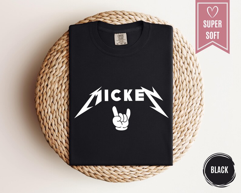 Pu&ograve; includere: T-shirt nera con la scritta "MICKEY" in bianco, con un carattere stilizzato, e un gesto della mano bianco. La maglietta &egrave; piegata su una superficie circolare intrecciata. Uno striscione rosa con un cuore e le parole "SUPER SOFT" &egrave; in alto a destra.