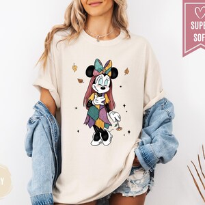 Puede incluir: Camiseta de color marfil con un personaje de dibujos animados de Minnie Mouse con un vestido y lazo de patchwork. El dise&ntilde;o incluye hojas que caen y el texto "SUPER SOFT" en una pancarta rosa. La camiseta es parte de una colecci&oacute;n de ropa.