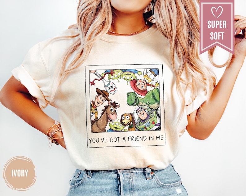Op de afbeelding: Ivoor kleurig T-shirt met een ingelijste afbeelding van Toy Story-personages, waaronder Woody en Buzz Lightyear. De tekst "YOU'VE GOT A FRIEND IN ME" staat onder de afbeelding. Een roze banner leest "SUPER SOFT".