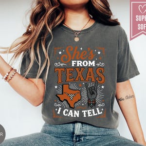 May include: A charcoal gray short-sleeve t-shirt featuring a graphic print with the text She's FROM TEXAS I CAN TELL. The design includes a silhouette of the state of Texas with a longhorn steer head, a hand making a rock sign, and stars.