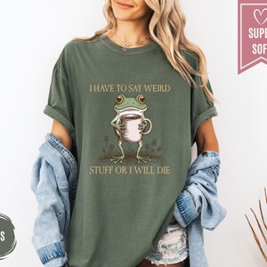 May include: A moss green t-shirt with the text "I HAVE TO SAY WEIRD STUFF OR I WILL DIE." The shirt features a cartoon frog holding a coffee cup. The shirt is made of a soft material.