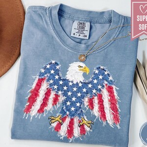May include: A blue jean Comfort Colors t-shirt with a patriotic eagle design. The eagle's wings are made of red and white stripes and blue stars. The shirt has a round neckline and a small tag. A gold necklace is draped across the shirt.