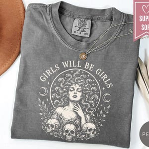 May include: A charcoal gray short-sleeve t-shirt featuring a white graphic print of a figure with snakes for hair, surrounded by crescent moons, leaves, and three skulls. The text "GIRLS WILL BE GIRLS" is arched above the central illustration.
