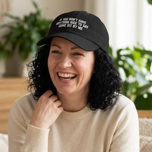 May include: Black baseball cap with white text: "IF YOU DON'T HAVE ANYTHING NICE TO SAY COME SIT BY ME." A person is wearing the cap and smiling. Green plants are in the background.