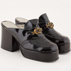 May include: Pair of black platform mules with a glossy finish. The shoes have a square toe, chunky heel, and a gold-coloured decorative accent. The platform height is approximately 5 cm, and the heel height is approximately 10 cm.