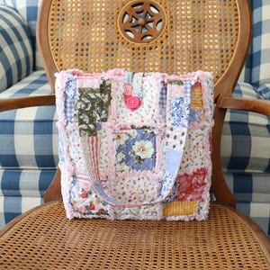 May include: A patchwork tote bag with a variety of floral and patterned fabrics in pink, blue, green, and white. The bag has a pink button closure and matching handles. The bag is sitting on a wooden chair.