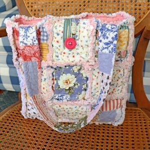 May include: A patchwork tote bag with a variety of floral and patterned fabrics in shades of pink, blue, and white. The bag features a large pink button closure and a ruffled edge. The bag is sitting on a wicker chair.