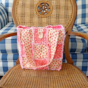 May include: A handmade tote bag with a patchwork design. The bag features a floral pattern in shades of red and cream, with pink trim and a button closure. The bag is sitting on a wooden chair.