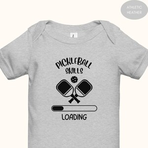 May include: Athletic heather gray baby bodysuit with the words "Pickleball Skills" above a graphic of two crossed pickleball paddles and a ball. Below the paddles is the word "Loading" with a progress bar.