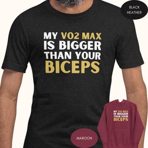 May include: A black heather t-shirt with the text "MY VO2 MAX IS BIGGER THAN YOUR BICEPS" in white and gold lettering. The shirt is shown on a model. A maroon hoodie with the same text is shown in the lower right corner.