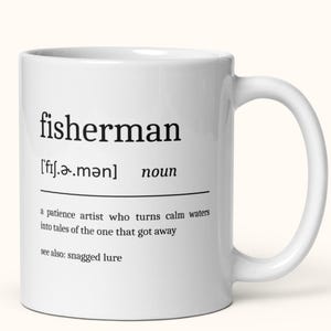 May include: White ceramic mug featuring the word "fisherman" in black, with its phonetic spelling and the word "noun." The definition reads: "a patience artist who turns calm waters into tales of the one that got away."