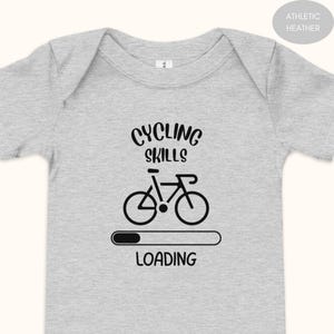 May include: Grey baby bodysuit with the text "CYCLING SKILLS LOADING" and a black bicycle graphic. The design includes a loading bar below the bicycle. The bodysuit is made of a soft, comfortable material, perfect for a baby.