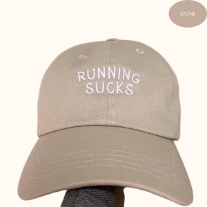 Può includere: Un cappello da baseball color pietra con il testo "RUNNING SUCKS" ricamato in bianco sul davanti.