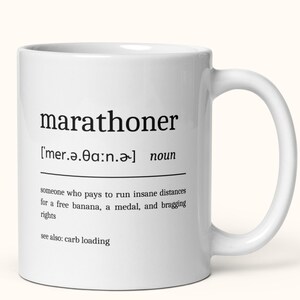Può includere: Una tazza da caffè in ceramica bianca con una definizione in bianco e nero della parola "maratoneta" stampata su di essa. La definizione recita: "['mer.ǝ.a:n.ǝ] nome qualcuno che paga per correre distanze folli per una banana gratis, una medaglia e diritti di vantarsi vedi anche: carico di carboidrati"