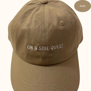May include: Khaki baseball cap with white embroidery that reads "On a Side Quest".